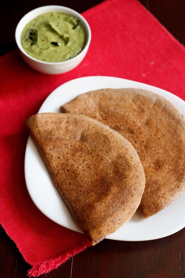 ragi dosa served on a white square plate on a red napkin with a side white bowl of green coconut chutney.
