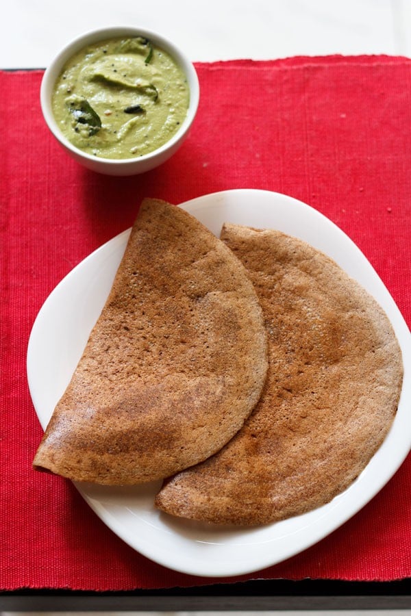 fermented ragi dosa served on a white square plate on a red napkin with a side white bowl of green coconut chutney.