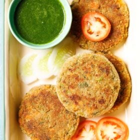veg cutlet placed on a tray with a few slices of tomatoes, cucumber and a light green bowl filled with green chutney