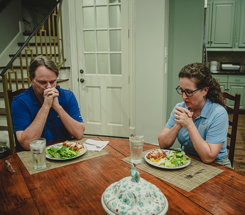 Mr. Kennedy and his wife, Gayle, pray together before dinner in their Houston home. Plates of salad and pasta sit on the table in front of them.