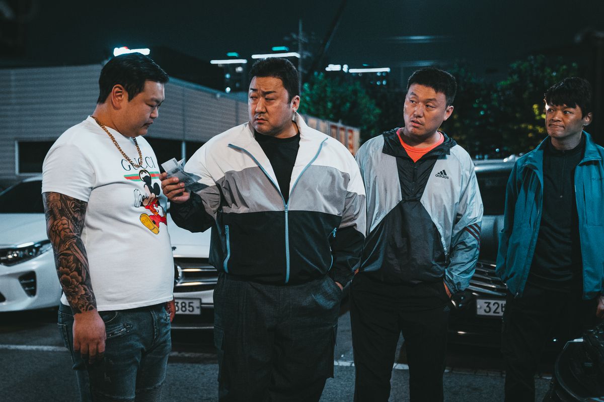 Ma Dong-seok, wearing a tracksuit stands next to another burly man in a white tee shirt and two of his fellow cops in The Roundup: No Way Out. He holds out a few pieces of paper bills to the man in the white tee.