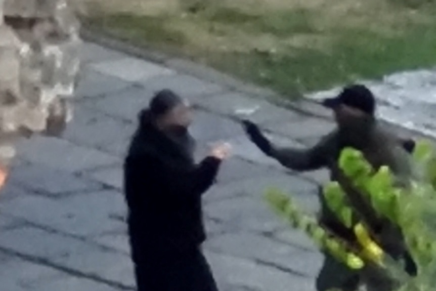 A blurry, zoomed-in aerial photograph shows a man with his face covered gesturing towards a man with a beard in a courtyard.