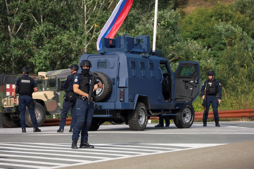 Police officers holding rifles and wearing face coverings stand on a crossroads near a blue armoured vehicle.
