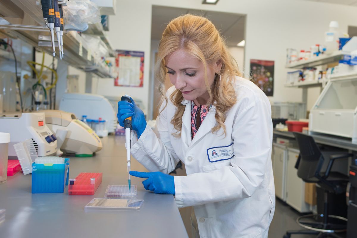 Melissa Herbst-Kralovetz wears a white lab coat and blue gloves while pipetting in her lab.