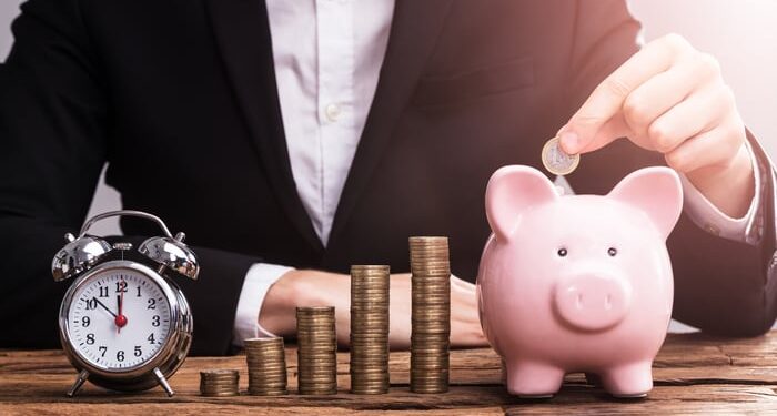 A person dropping a coin into a piggy bank, beside which is an alarm clock and ascending stacks of coins.