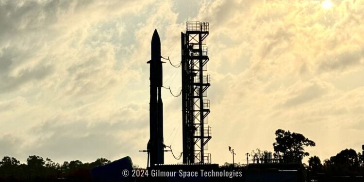 the silhouette of a rocket stands next to a launch tower with staircase, against a bright cloudy sky.