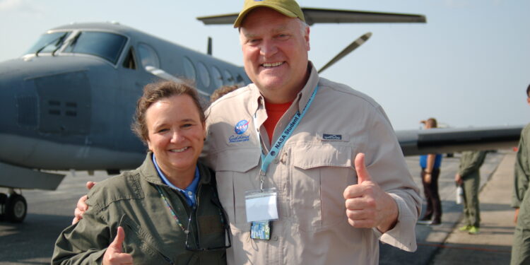 Woman wearing a green jacket and man wearing a tan button down shirt with the Goddard logo stand on a runway in front of a plane.