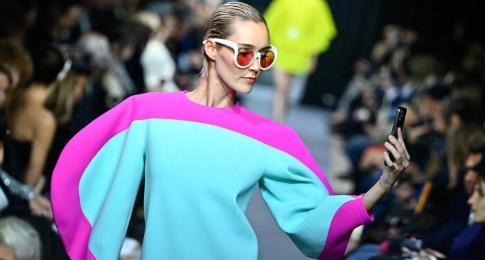 A model showcases a vibrant creation by Pierre Cardin, featuring a bold pink and turquoise ensemble. She wears oversized sunglasses and holds a smartphone while walking the runway