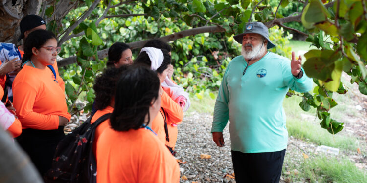 A Puerto Rican man with a bushy white beard, green bucket hat, teal longsleeve shirt, and black shorts gestures to a group of high school students wearing neon orange shirts.