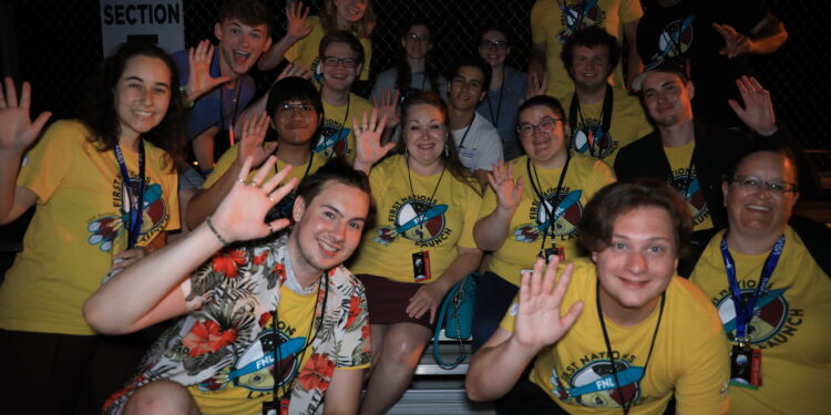 A group of people wearing yellow t-shirts wave at the camera as they sit outside at night. They are members or advisors of winning teams from the 14th First Nations Launch High-Power Rocket Competition. The competition comprises students from tribal colleges and universities, Native American-Serving Nontribal Institutions, and collegiate chapters of the American Indian Science and Engineering Society.