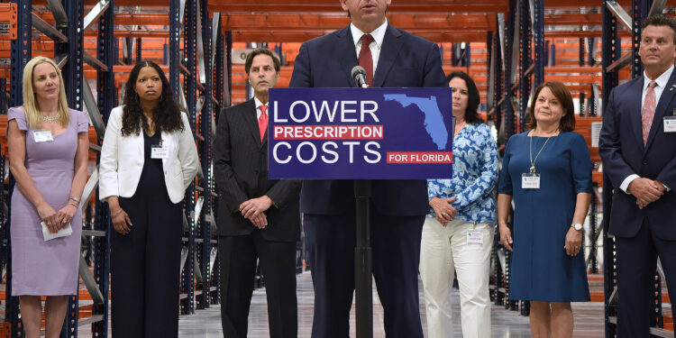 A photo of Ron DeSantis speaking inside a warehouse at a podium with a sign that reads, "Lower drug costs."