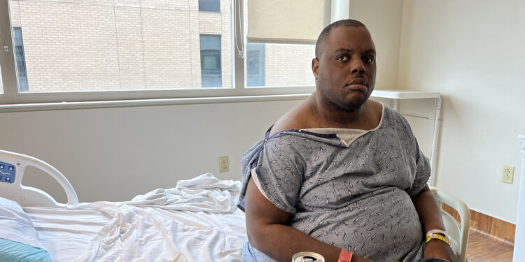 A man wearing a hospital gown is sitting on a bed in a hospital room alone.