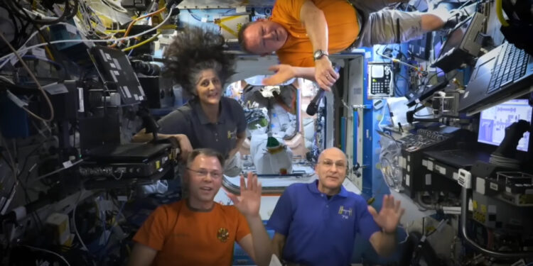 three men and one woman wave from inside the international space station