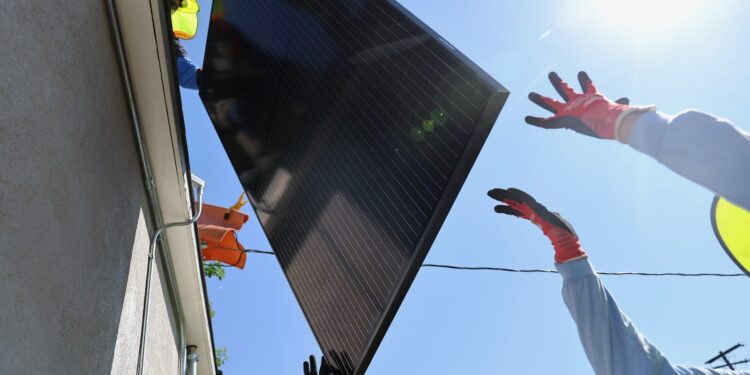 Two workers hand a solar panel to another worker on the roof a house.