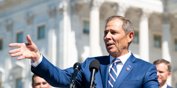 Photo of a man in a blue suit gesturing in front of a microphone in front of the Capitol