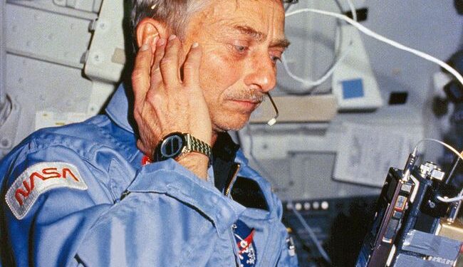 a man in a light blue flight set listens to a radio in a cramped room filed with wires and computers