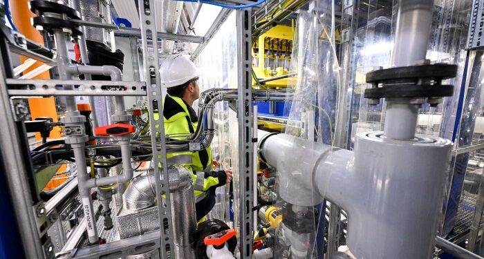 An employee wearing a hard hat and high-visibility jacket examines equipment surrounded by pipes and machinery at an energy lithium electrolysis optimisation plant in Germany.