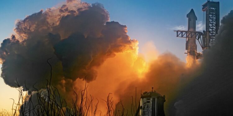 A chrome finned rocket lifts off next to a dark grey launch tower with black extended arms. Smoke billows up, obscuring all but the rocket's second stage, glowing orange and rising into the clue blue sky. In the foreground, grass reeds and an old test launch vehicle in the distance.