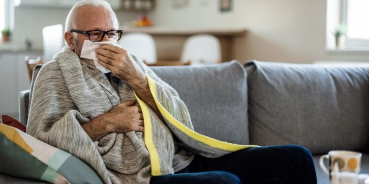 Elderly man catches a sneeze in a tissue, sitting on a grey sofa