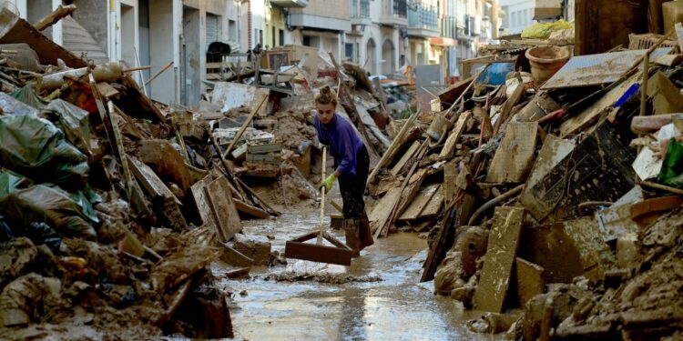 a woman in a purple shirt uses a broom to push away debris in a street full of debris on either side. she is ankle-deep in water. There is light coming from behind her. The street is narrow.