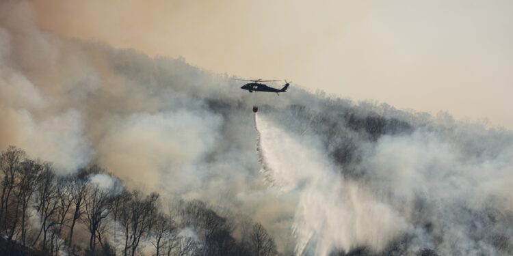 A helicopter sprays a liquid over a forest engulfed by smoke