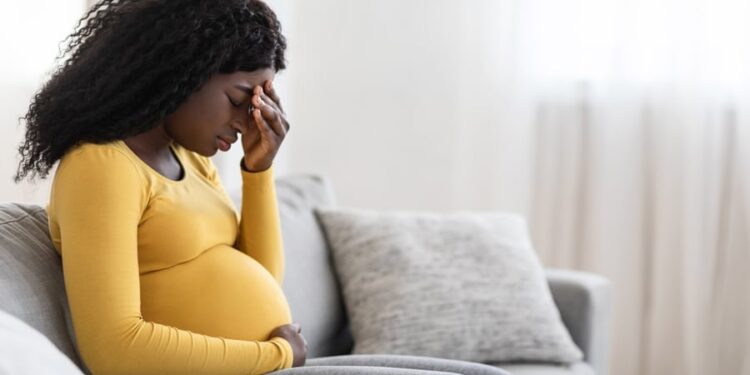 Young pregnant black woman suffering from headache or migraine, feeling sick, sitting on sofa at home, free space. Sad african american expecting lady touching her forehead and big tummy, home alone