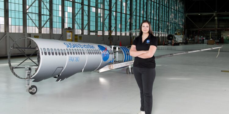 A woman wearing a black polo shirt with the NASA insignia, dark gray pants, and white shoes stands inside an aircraft hangar in front of a scaled aircraft model. The words â€œSUSAN Electrofanâ€ and â€œPAX 180â€ are printed on a graphic along with fuselage, with a large NASA insignia in the center of the plane. The rear of the aircraft model, along with the wings and tail, are taken apart to display the inner components that make up its electrified propulsion system.