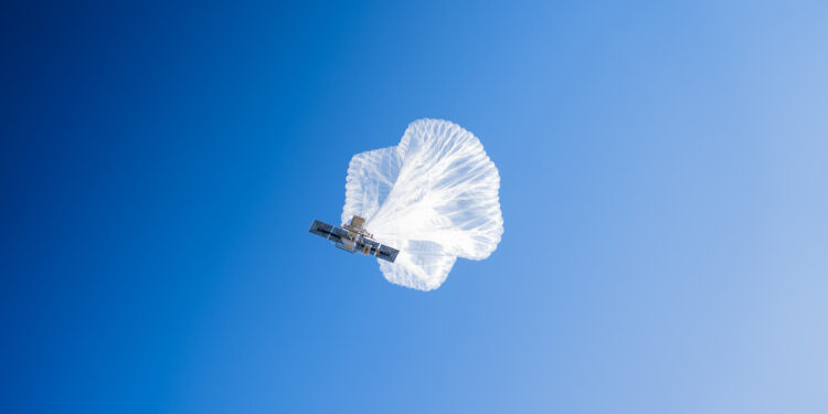 A high-altitude balloon is photographed from below as it ascends into a blue sky. The white balloon is partially inflated and the cell transmitter and other equipment is visible.