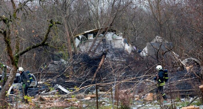 Emergency workers in protective gear examine debris at the site where a DHL cargo plane crashed into a house near Vilnius, Lithuania. The area is strewn with wreckage and fallen trees.