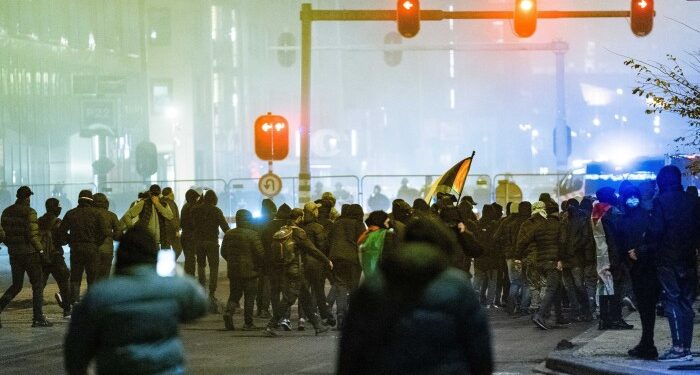 Protesters, many wearing dark clothing and masks, gather in the street during a pro-Palestinian demonstration in Amsterdam. Some carry flags, with a backdrop of urban lighting and visible police presence