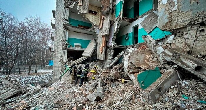 Rescue workers are clearing rubble from a severely damaged residential building. The structure is partially collapsed, with debris scattered around