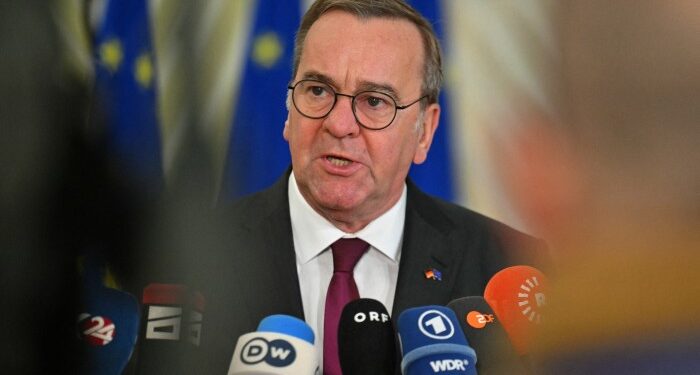 German Defence Minister Boris Pistorius speaks to the press, surrounded by microphones from various media outlets. He is wearing a dark suit and a maroon tie, standing in front of a backdrop featuring EU flags.