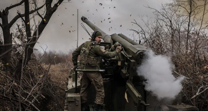 A Ukrainian serviceman operates a 2S5 152 mm self-propelled howitzer, firing towards Russian positions. The scene is set in a wooded area, with smoke and leaves visible around the artillery.