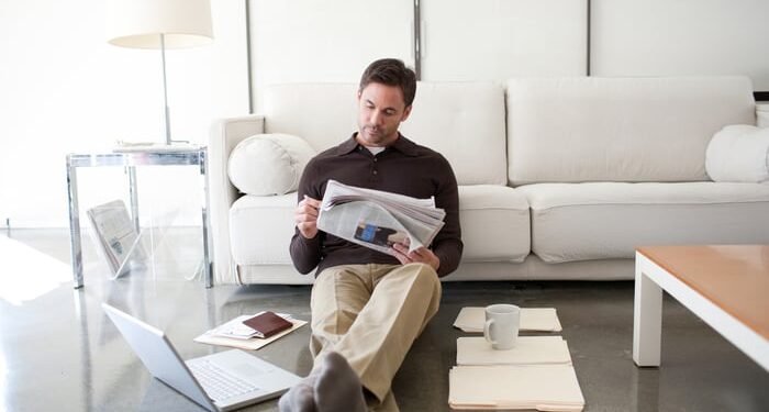 An investor sitting against a couch while reading the newspaper, with a laptop and piles of manilla folders on the floor.