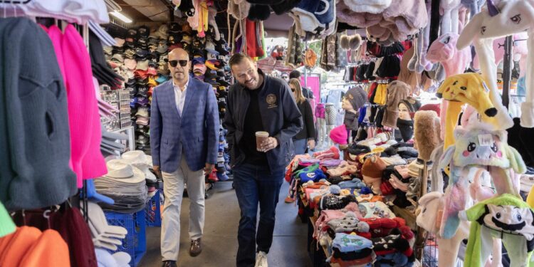 Two men walking between displays of caps, cowboy hats, novelty headwear and balaclavas.