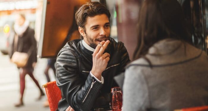 Person seated at and outdoor cafe holding a cigarette.