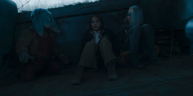 Still from Star Wars: Skeleton Crew TV series. Three children – Neel (blue elephant-like alien boy), Fern (human girl), and KB (girl with an asymmetrical white bob and wearing visor across her eyes) – are sitting with their backs against a wall in a dark room, hiding.