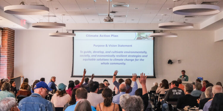 A man at a podium stands next to a projector screen with the words, "Climate Action Plan: Purpose & Vision Statement. To guide, develop, and cultivate environmentally, socially, and economically resilient strategies and equitable solutions to climate change for the whole community." There is a crowd of several dozen people in front of him. Some of their hands are raised.